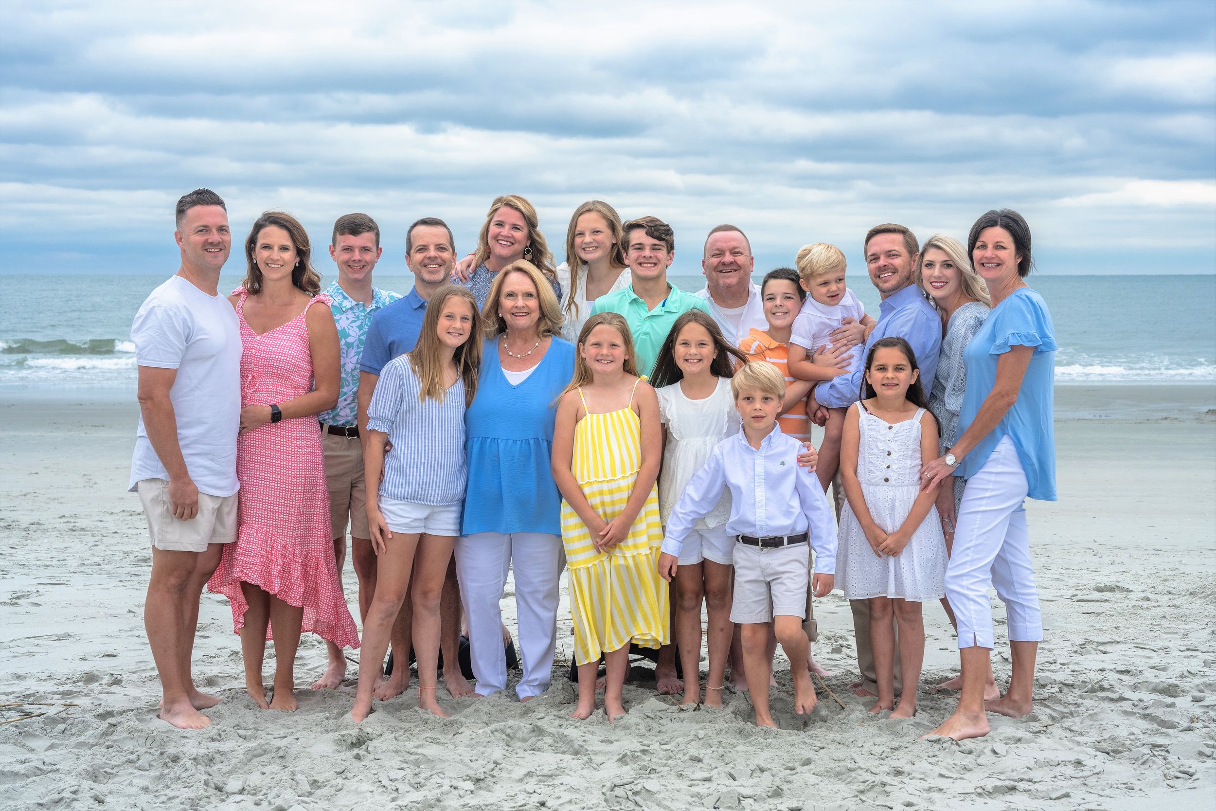 Large family group portrait on a sandy beach with overcast skies, dressed in coordinating pastel outfits.