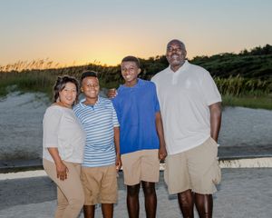A family of four poses together on a sandy beach at sunset with sea grass and dunes in the background.