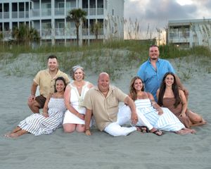 A group relaxes together on a sandy beach with dunes and beach houses visible in the background.