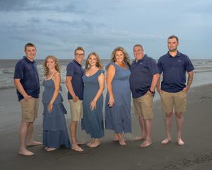 A group in coordinated blue and beige outfits poses together on a sandy beach at sunset.