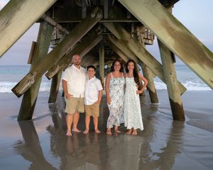 A group stands beneath a wooden pier on a beach during golden hour with ocean waves in the background.