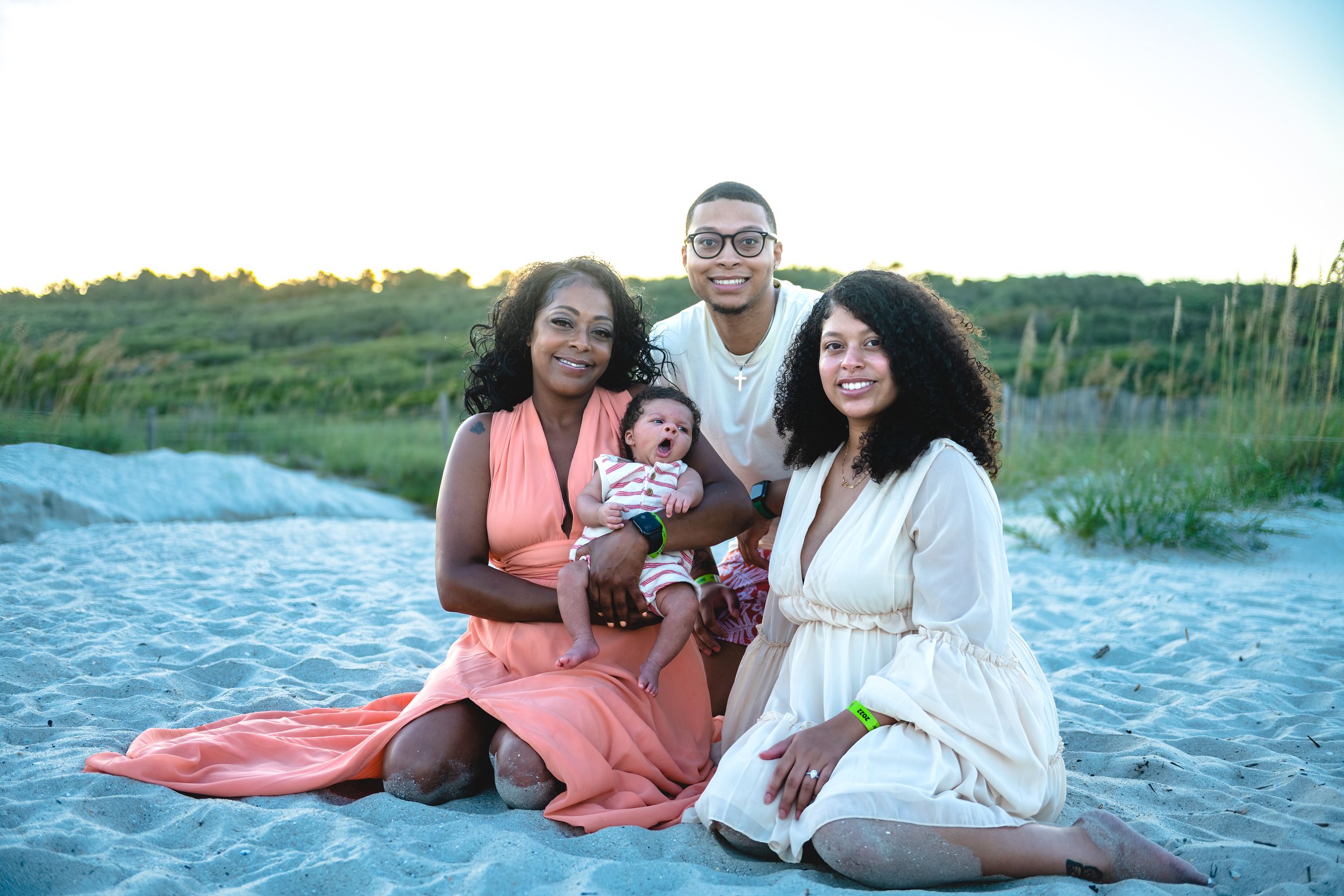 Family in coordinating coral and white outfits poses together on a sandy beach at sunset.