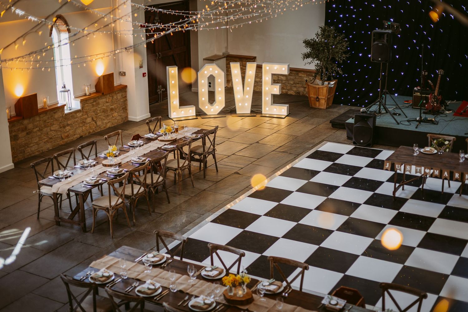 String lights illuminate a rustic wedding reception venue with black and white checkered dance floor and wooden tables.