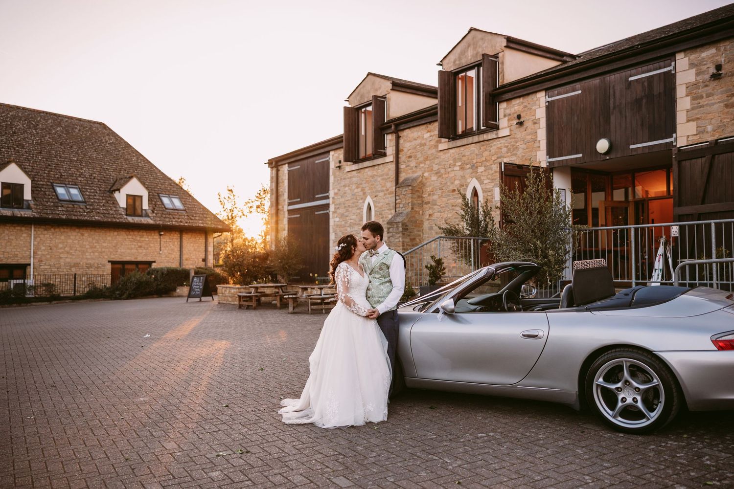 Newlyweds pose with a silver sports car outside a brick building during sunset.
