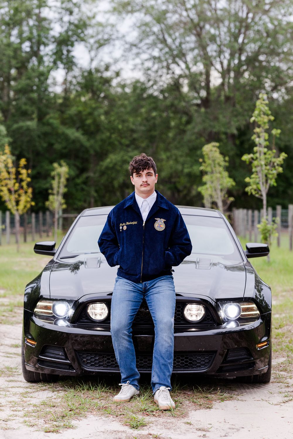Student in FFA jacket poses with black sports car in outdoor setting.