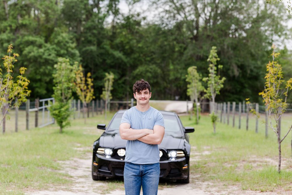 A black car parked in a grassy field with trees in the background and a person standing nearby.