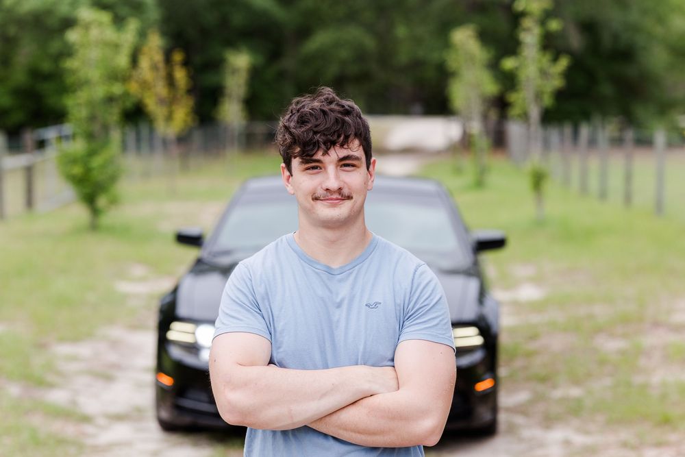 A person in a light blue t-shirt poses with crossed arms and a friendly smile in an outdoor setting.