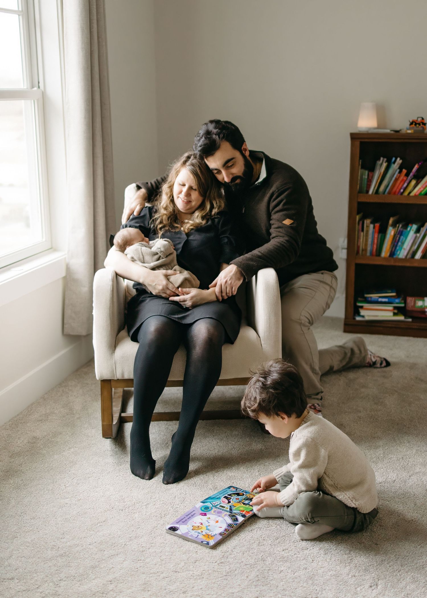 Family stands together by large window looking lovingly at their newborn baby in a tender indoor moment.