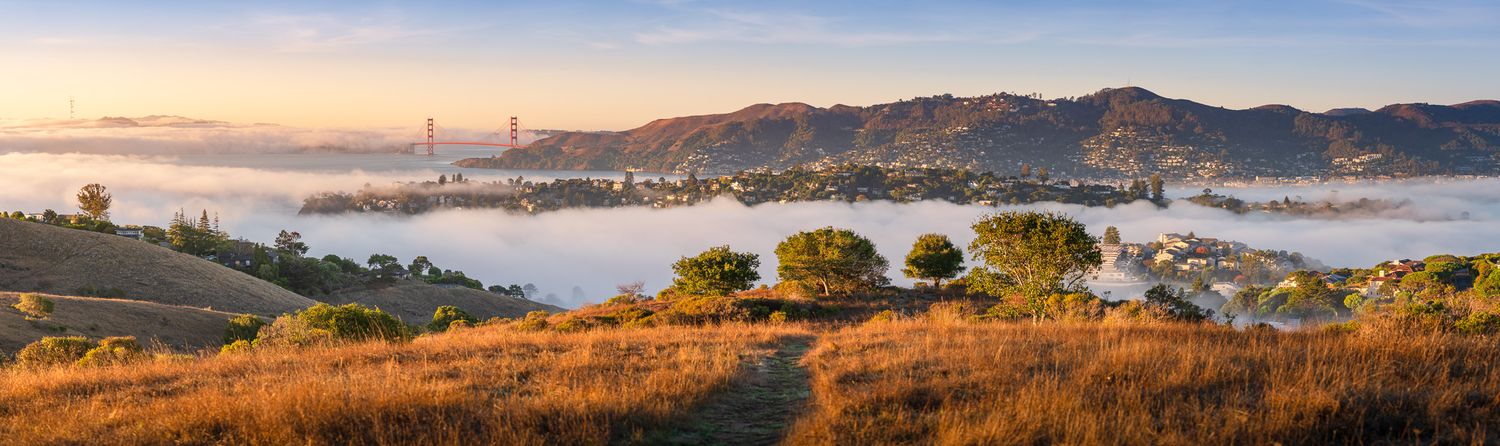 Fog rolls over the Golden Gate Bridge and San Francisco Bay during a colorful sunset with mountains in the background.