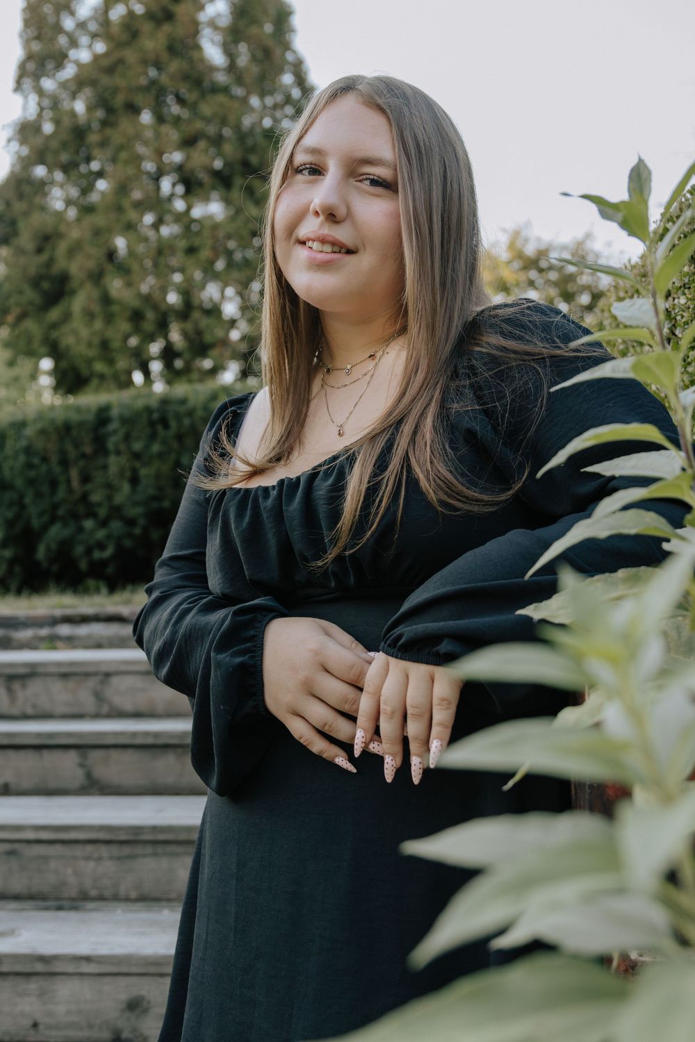 Someone poses in a black dress against foliage and brick architecture during a sunny outdoor shoot.