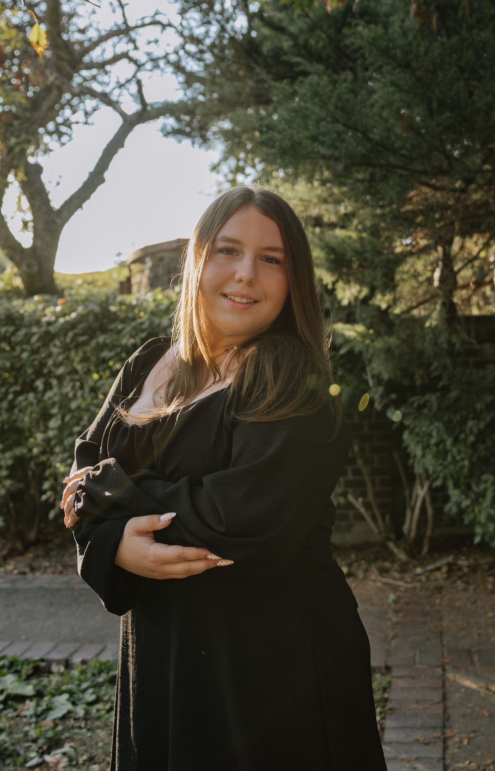 Someone in a black outfit poses expressively against a backdrop of trees and greenery.