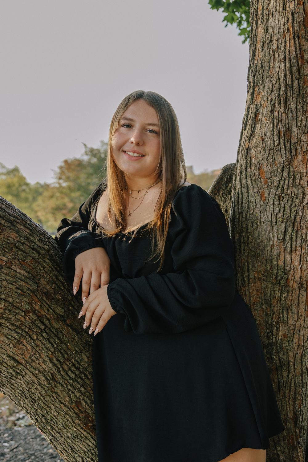 Portrait of someone in a black dress leaning against a tree trunk in an outdoor setting.