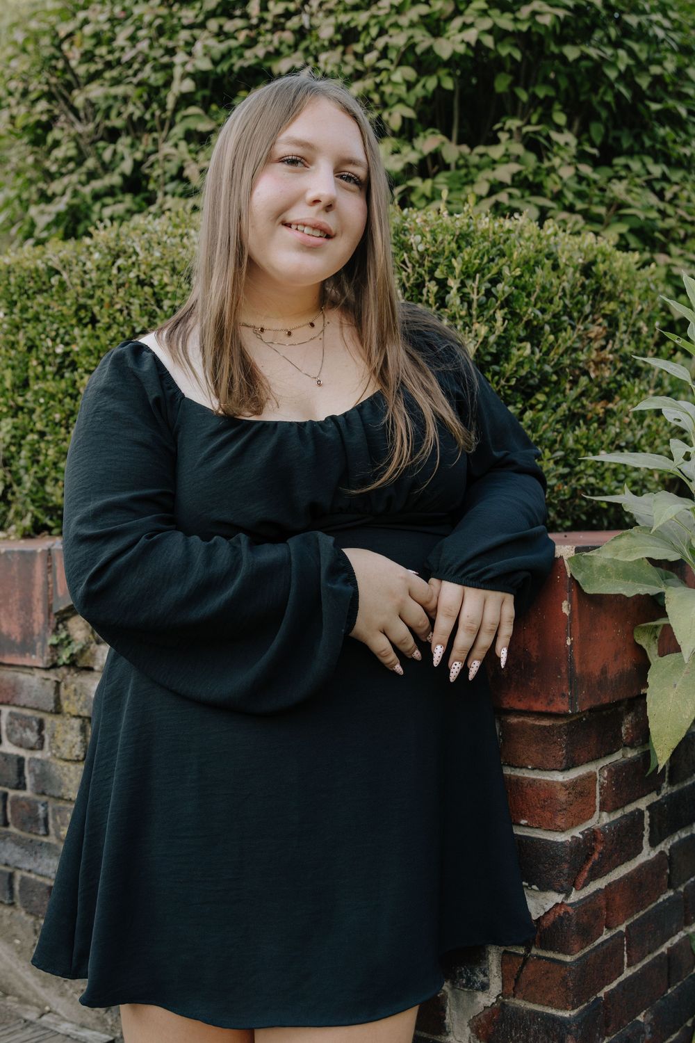 A person in a black long-sleeve top poses casually against a brick wall surrounded by greenery.
