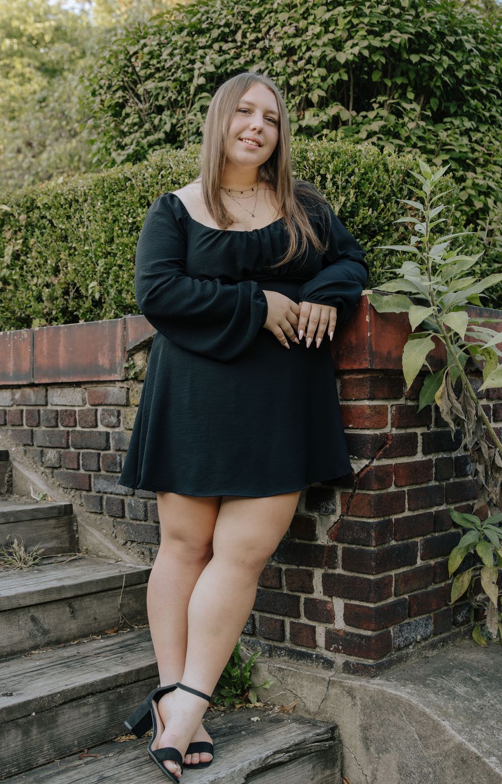 A person in a black dress sits on stone steps next to a brick wall in an outdoor garden setting.