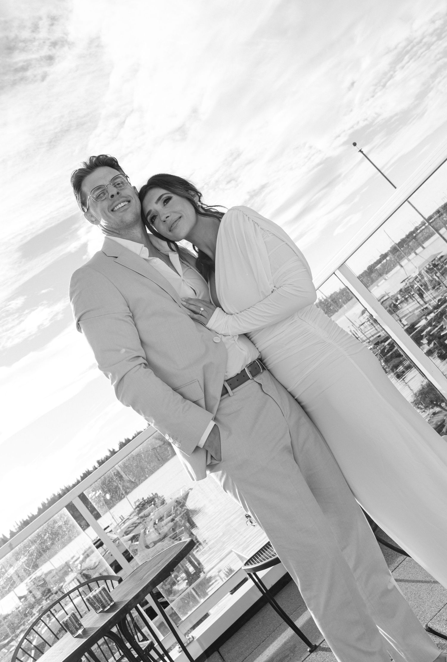 A black and white photograph of a couple in light colored attire embracing on a balcony overlooking a city.