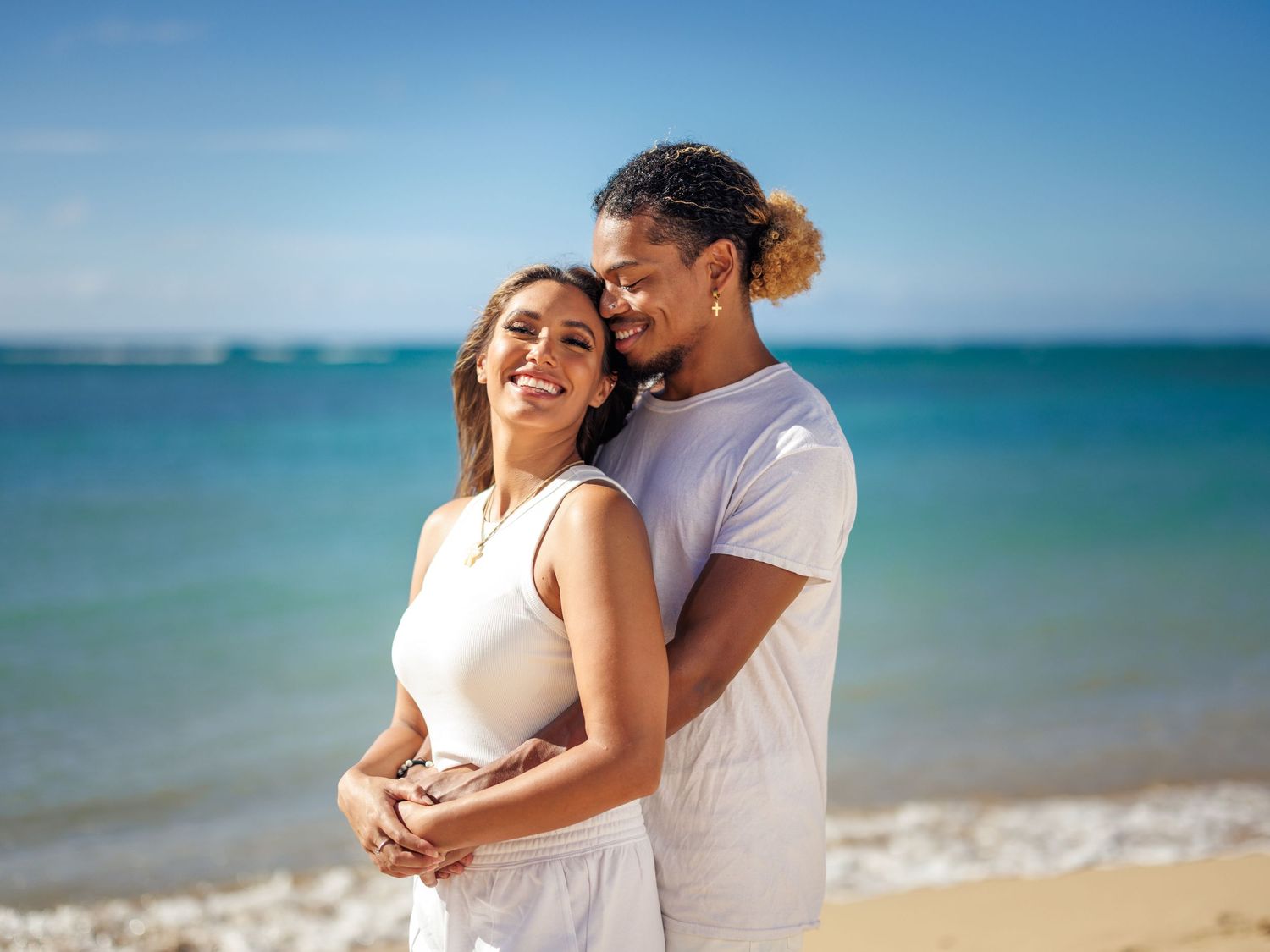 Happy couple embracing on a sunny tropical beach, wearing white outfits with turquoise ocean waves in the background.
