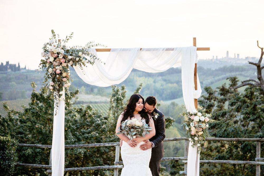 A romantic wedding ceremony overlooking rolling hills of San Gimignano with a floral arch and flowing white fabric draping.