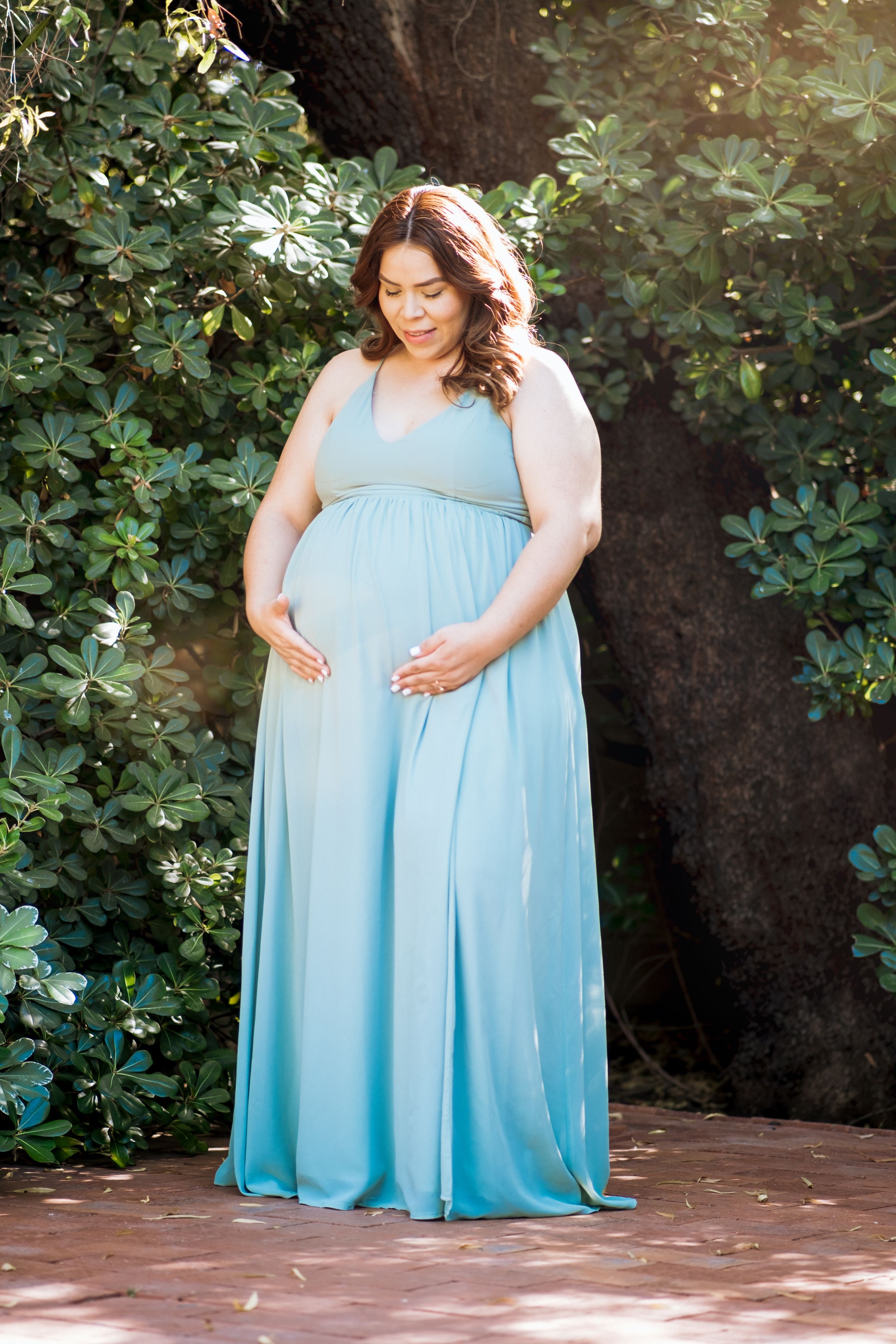 A pregnant woman in a flowing light blue dress poses in a garden setting.