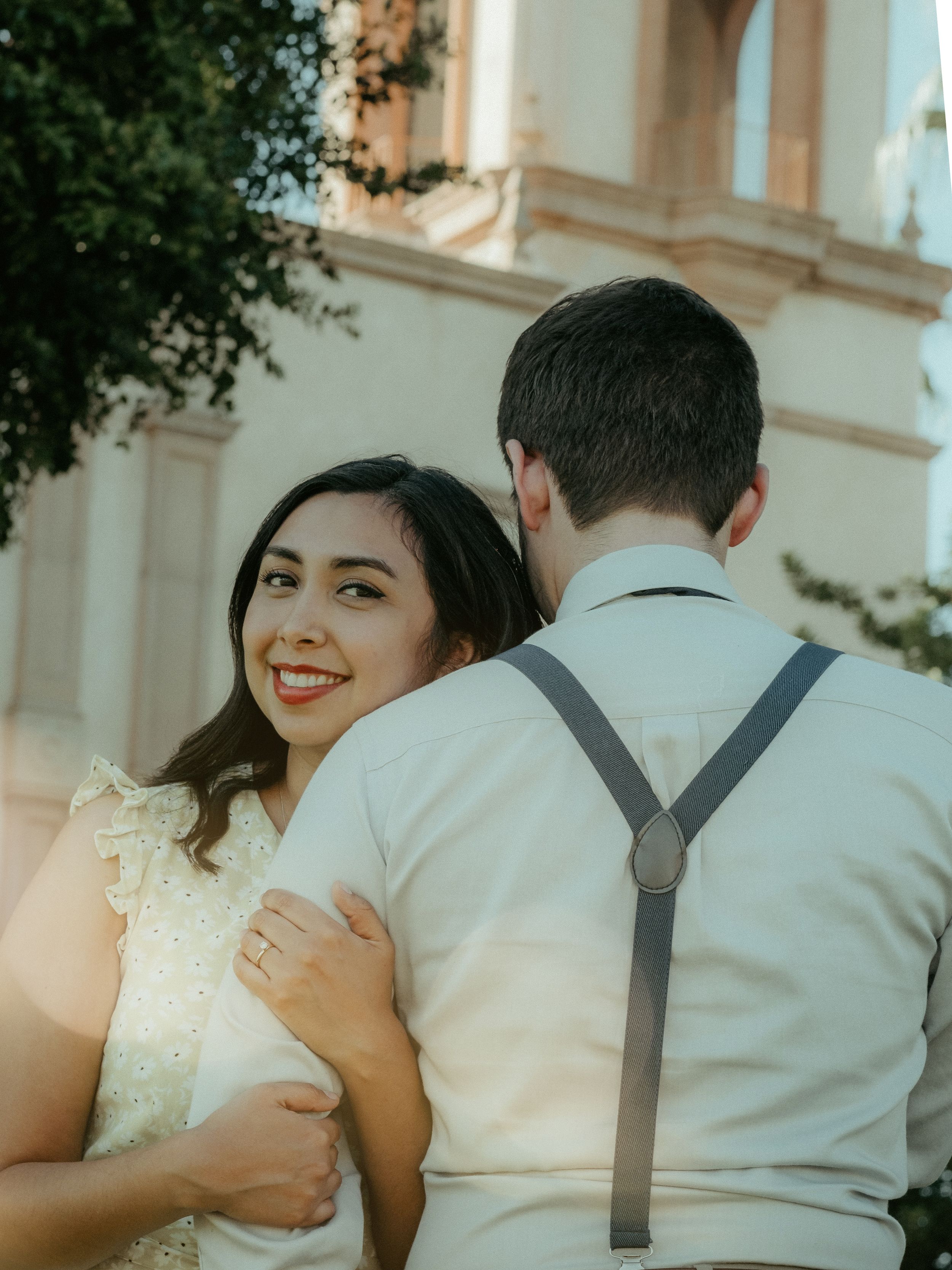 A bride in a lace dress smiles while standing next to her groom in suspenders outside a church.
