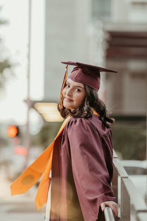 A graduate in maroon cap and gown poses with orange stoles against an urban backdrop.