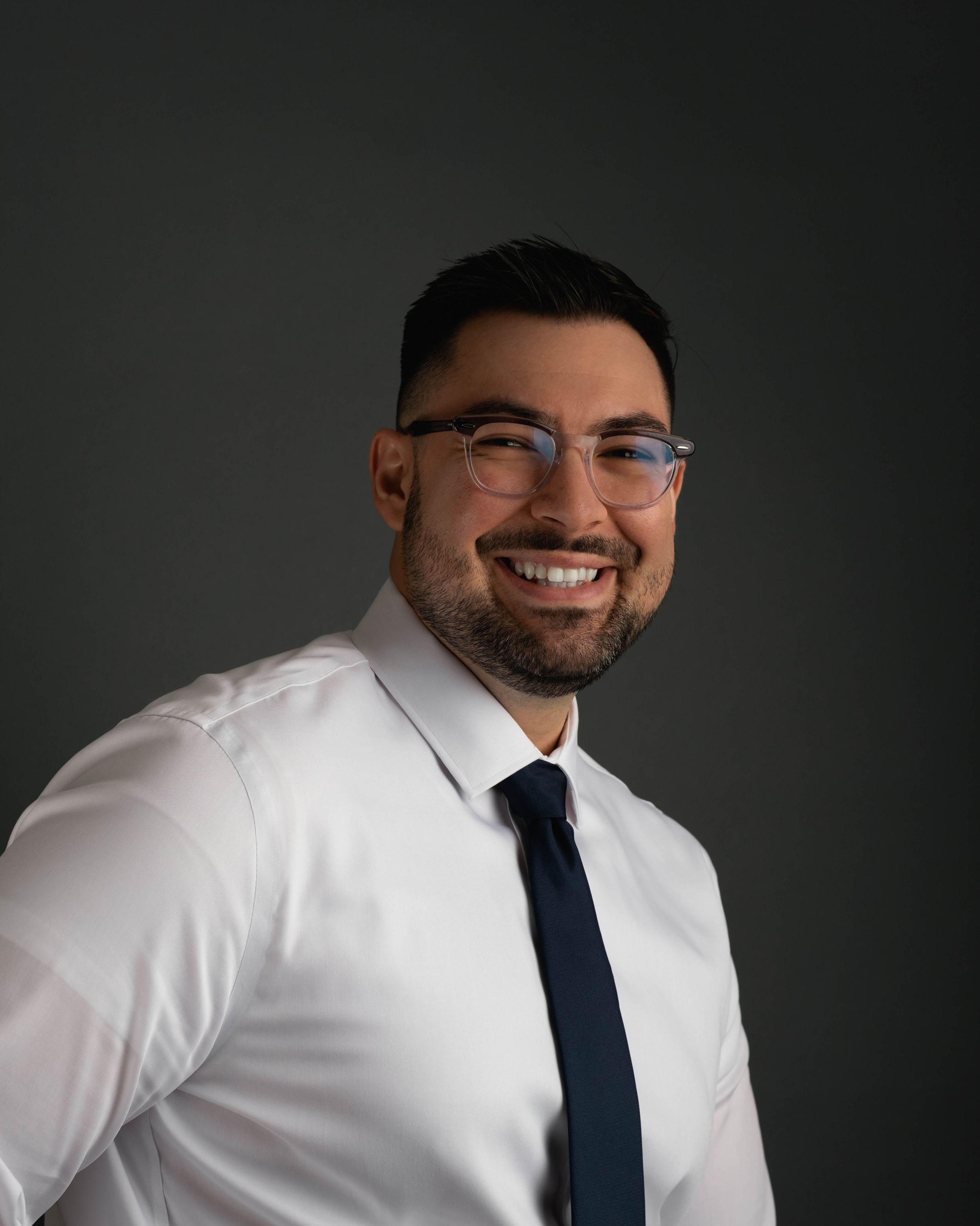 Professional headshot of someone in glasses, white shirt and navy tie against dark background.