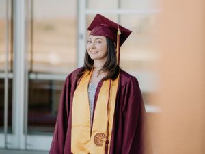 A graduate in maroon cap and gown with gold stole smiles in front of a modern building.