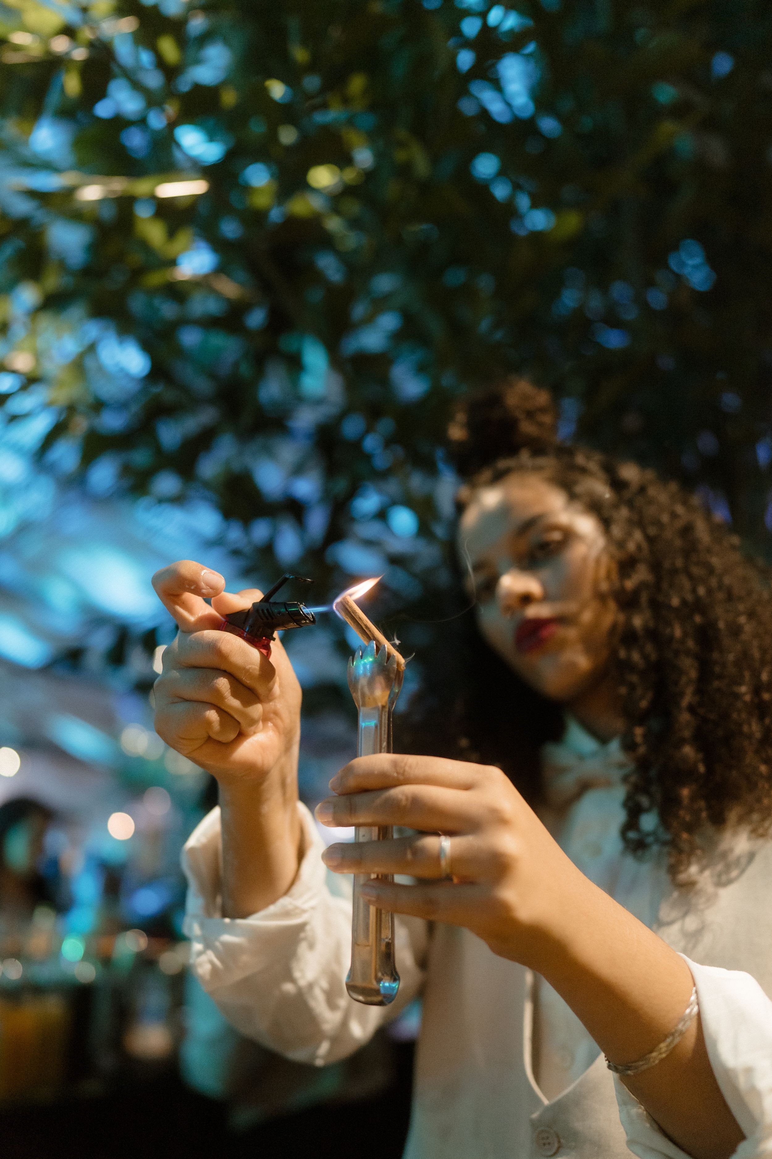Woman in white shirt lighting a candle against blurred bokeh background with foliage.
