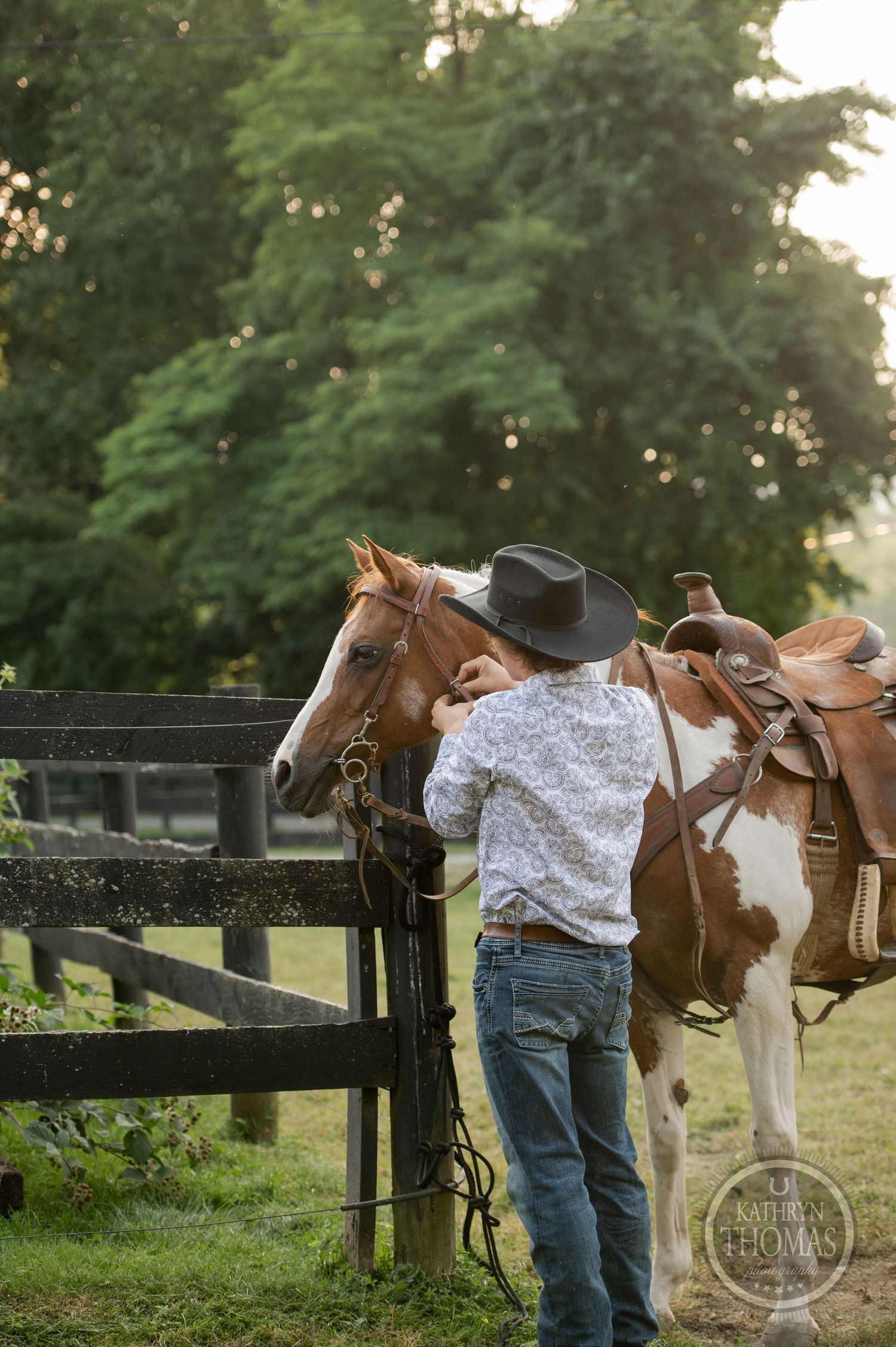 A western-dressed person tends to a saddled paint horse by a black fence at dusk.
