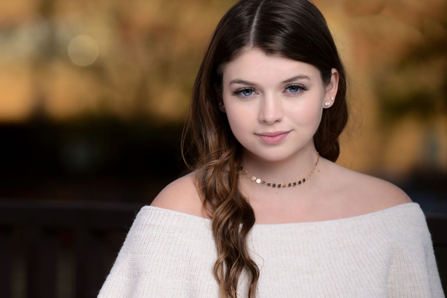 A figure in an off-shoulder white top with long wavy brown hair and a delicate necklace poses outdoors in soft lighting.