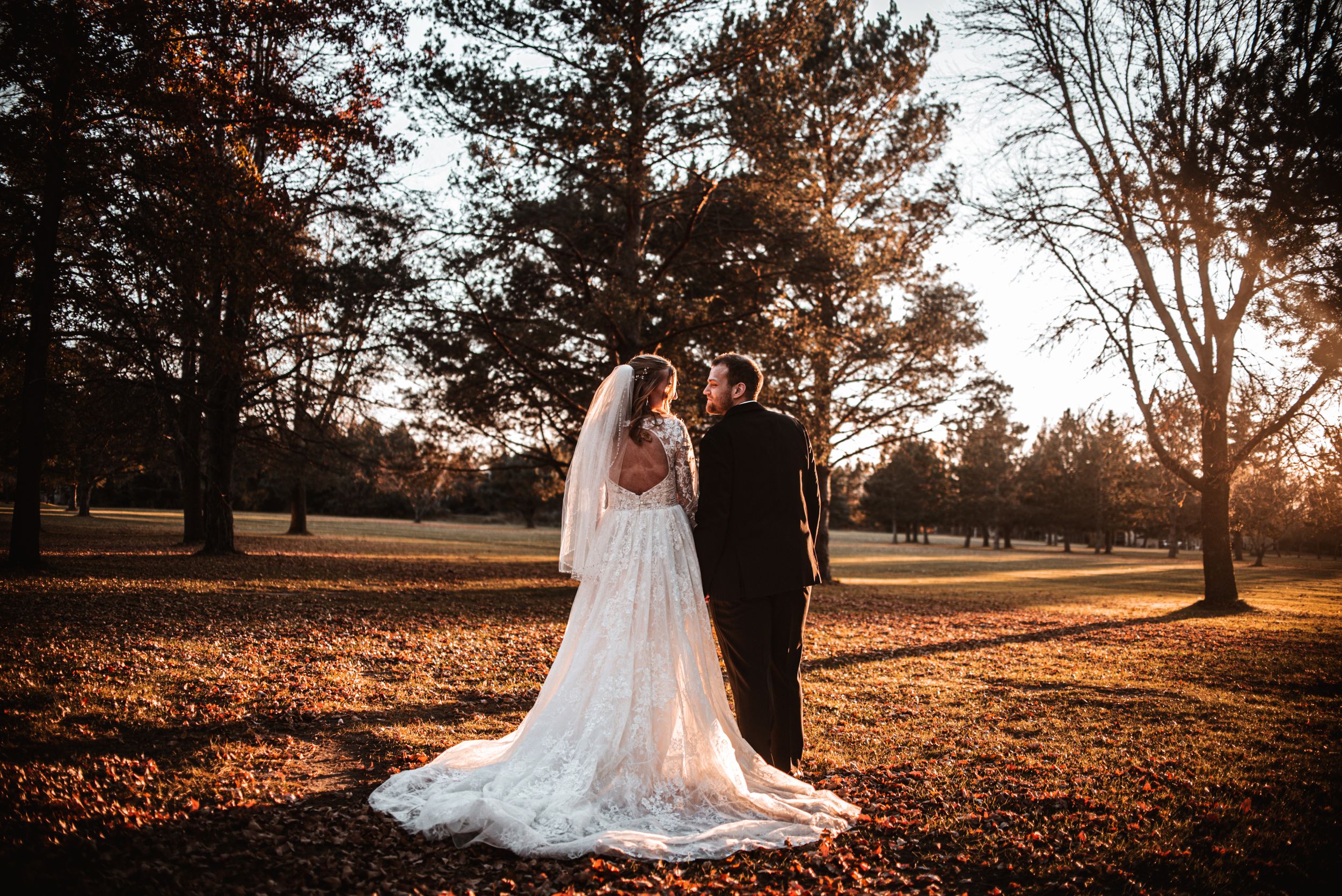 A couple embraces during sunset at a park with autumn leaves scattered on the ground as golden light filters through trees.