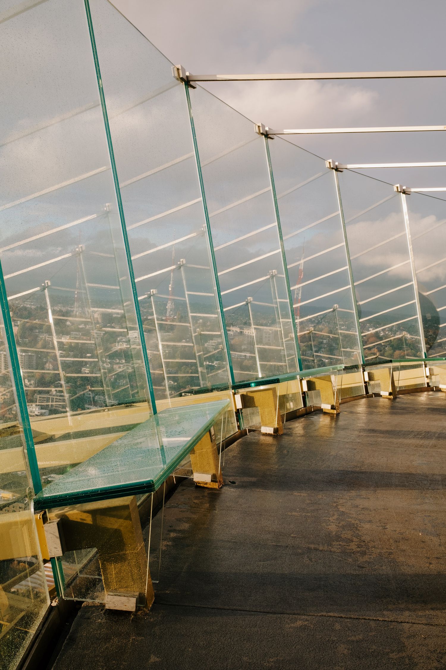 Modern glass-walled observation deck with curved benches and metal support beams overlooking a cityscape beneath cloudy skies.