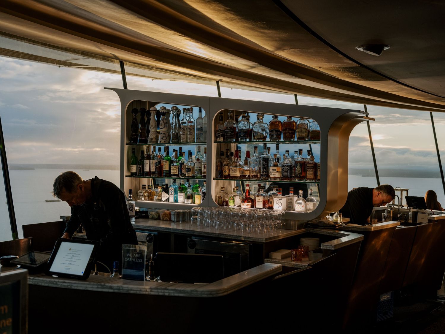 Elegant cruise ship bar with backlit liquor display and bartender working against a sunset ocean view through windows.