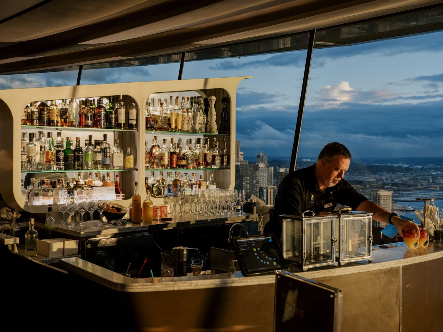 A bartender works behind a curved bar with illuminated shelves of liquor bottles and a scenic city view in the background.
