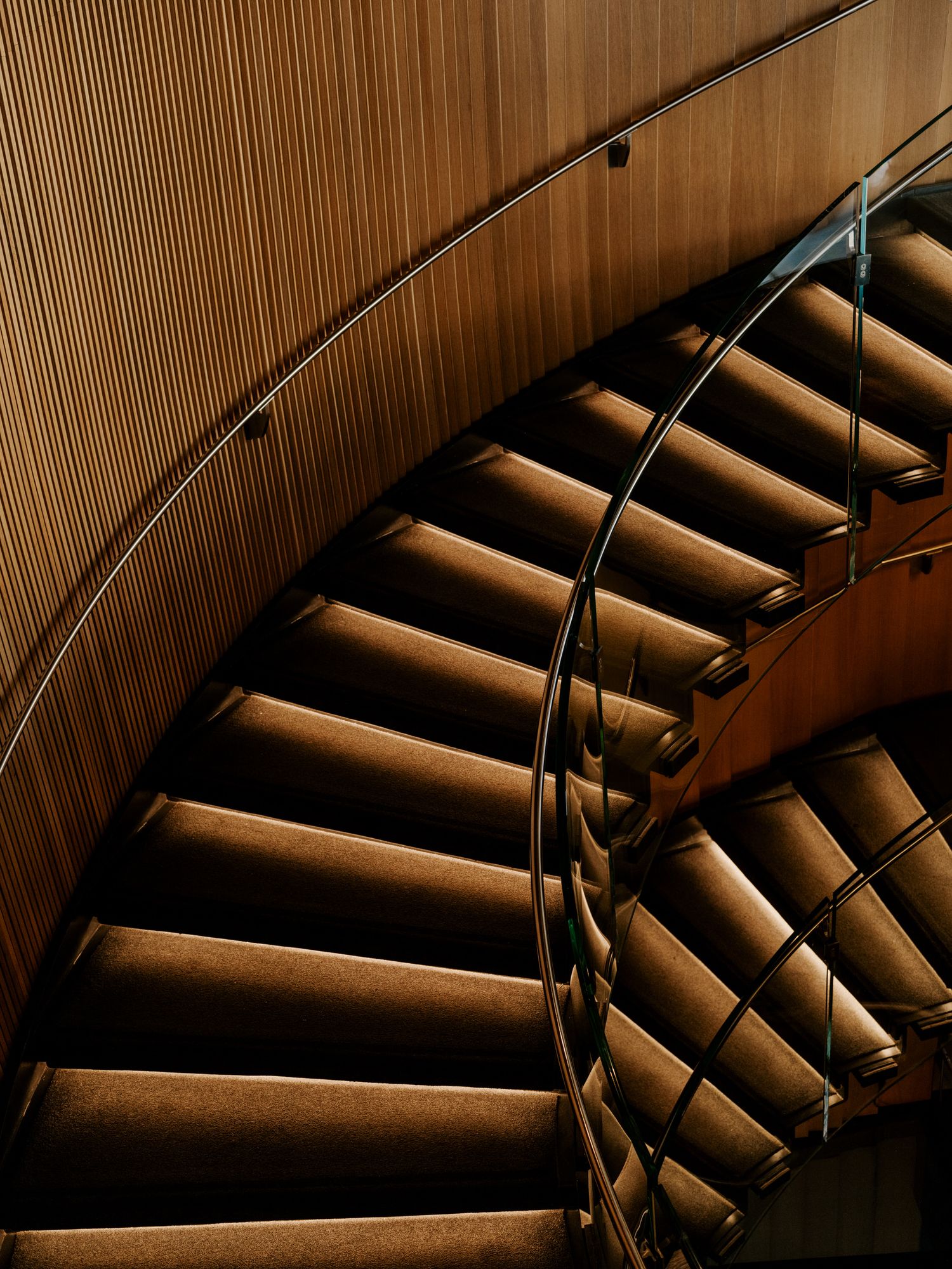 Modern curved wooden staircase with elegant handrail and wood paneled walls illuminated by warm lighting.