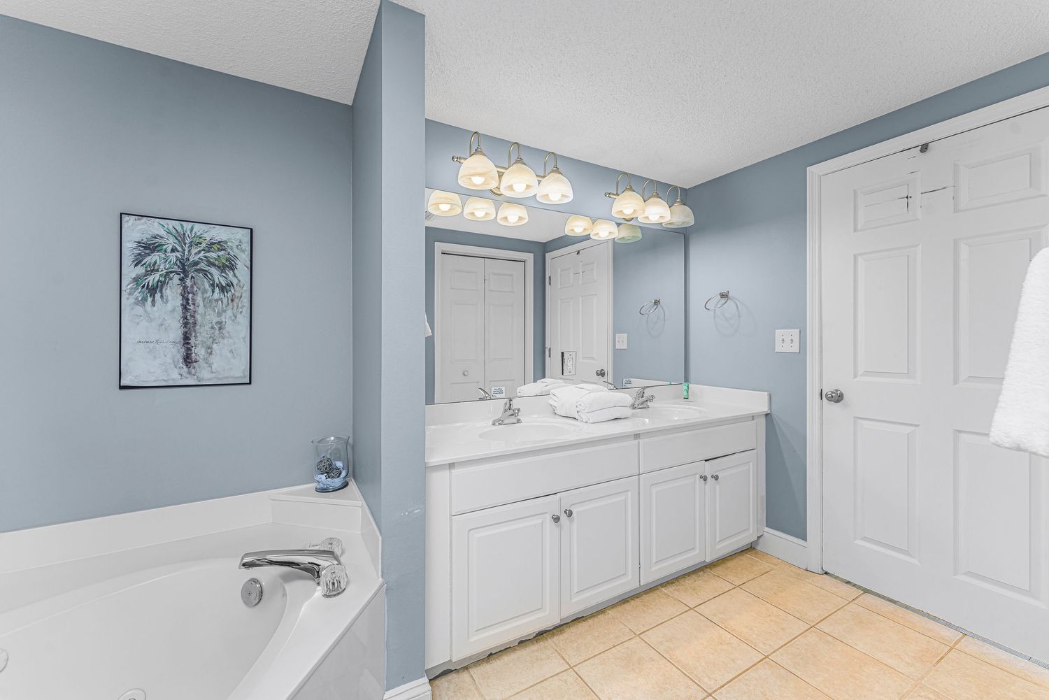 Modern master bathroom with light blue walls, white vanity cabinets, dual sinks, and beige tile flooring.