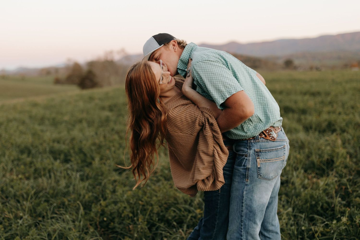 A romantic couple shares a passionate embrace in a field during sunset.