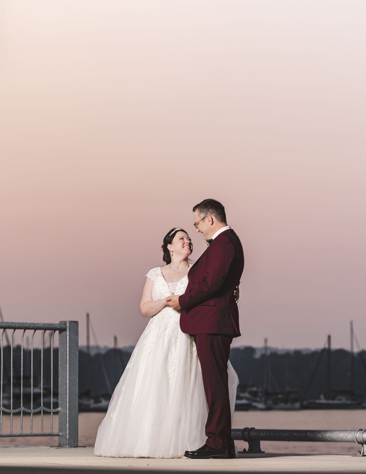 A couple in wedding attire shares an intimate moment at sunset on a waterfront balcony.
