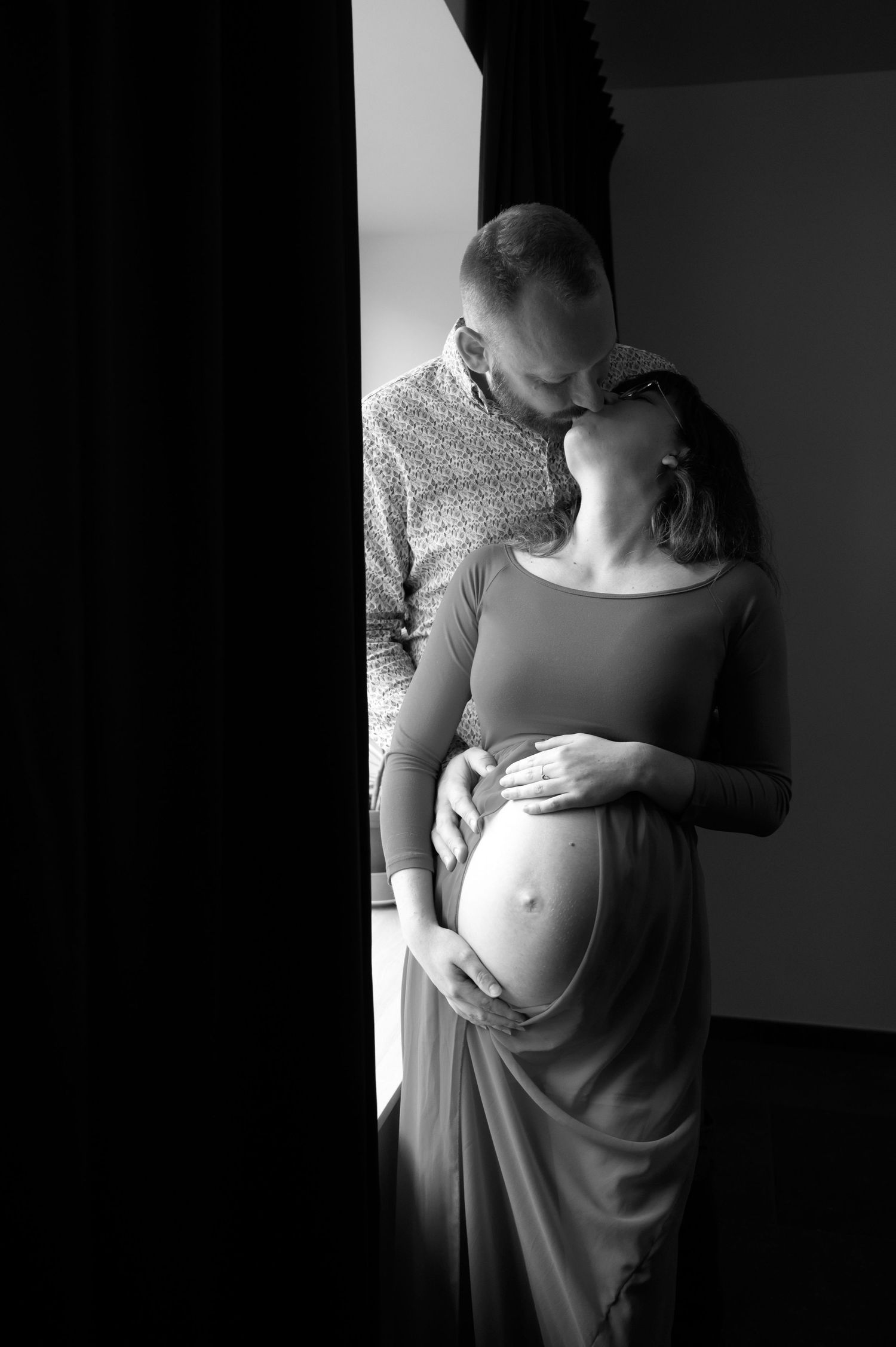 À Saint-Hubert, séance photo de grossesse en couple s’embrassant devant une fenêtre, ventre apparent, en noir et blanc