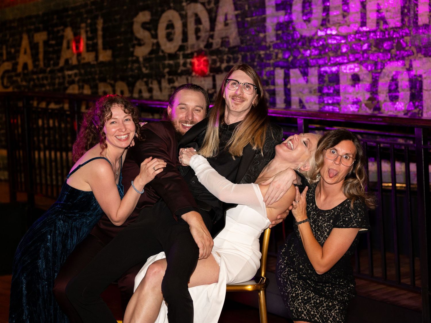 A group of friends pose and laugh together at a nightclub with neon lights and signage in the background.