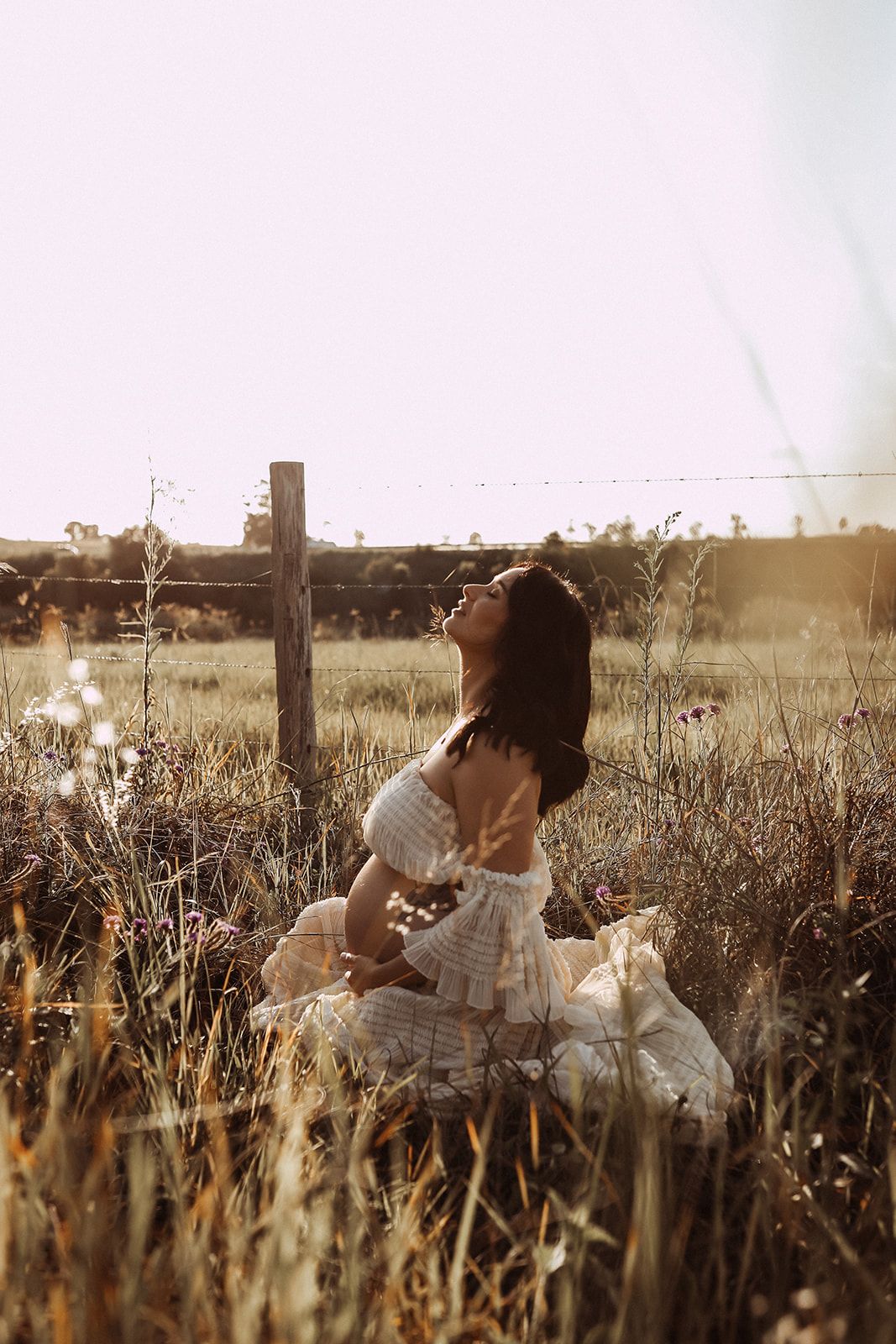 Woman in white dress sitting in tall grass during golden hour photoshoot.