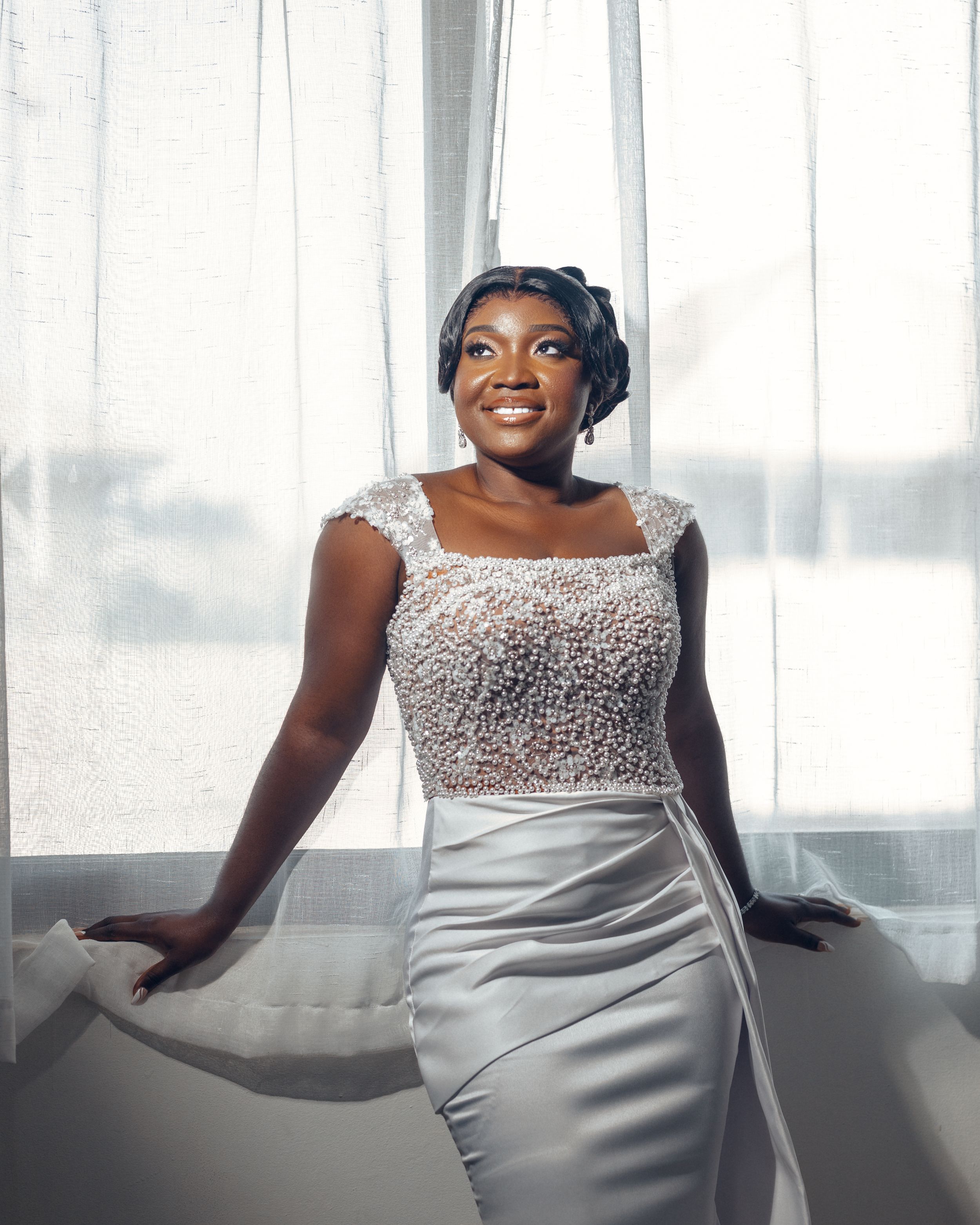 Bride in fitted white gown with beaded bodice poses by window in natural light.