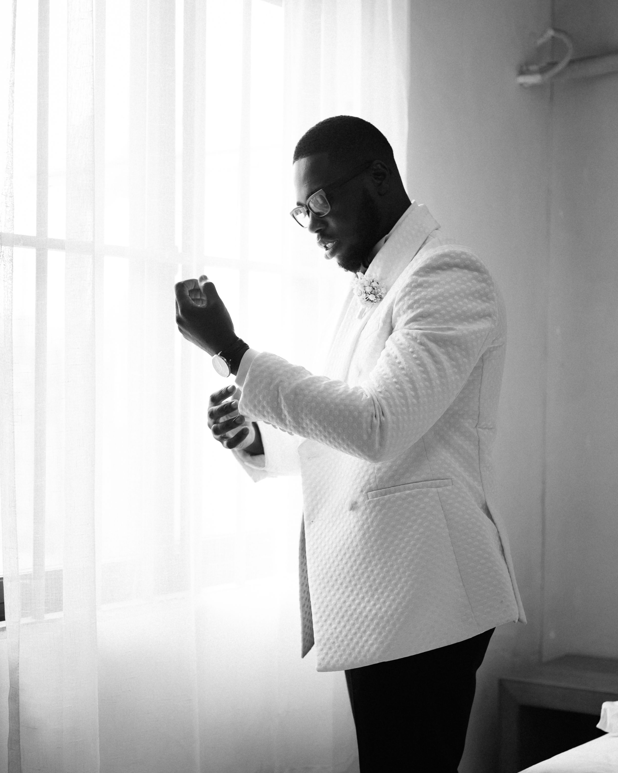 Groom in white textured jacket adjusts cufflinks by window.