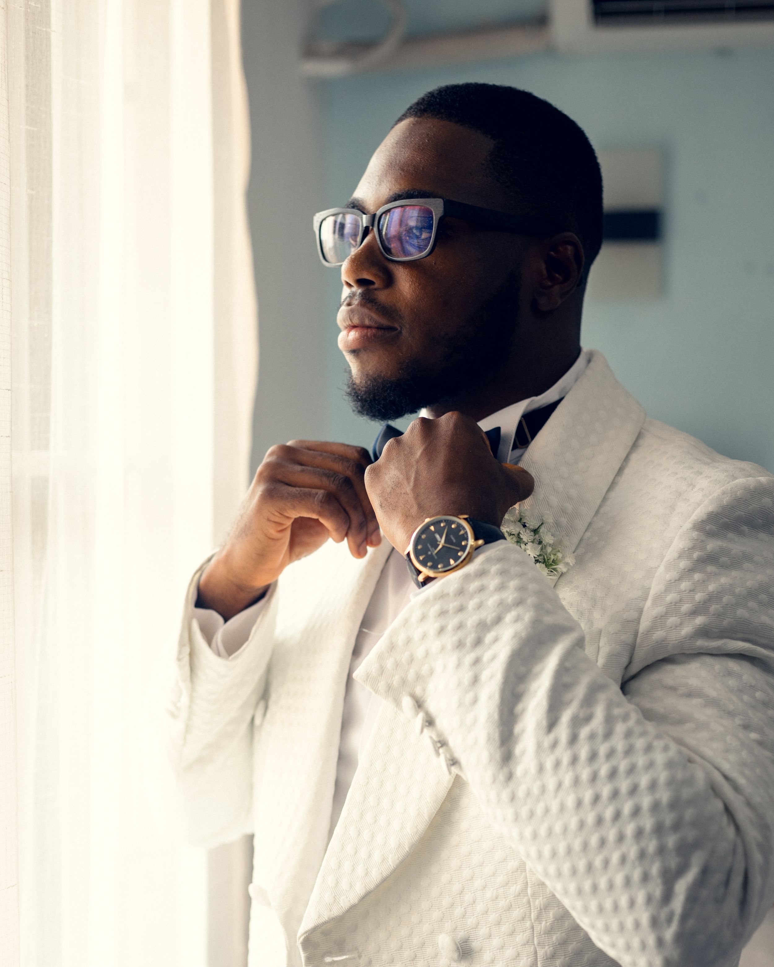 Groom wearing white quilted dinner jacket and glasses adjusts bowtie near window.