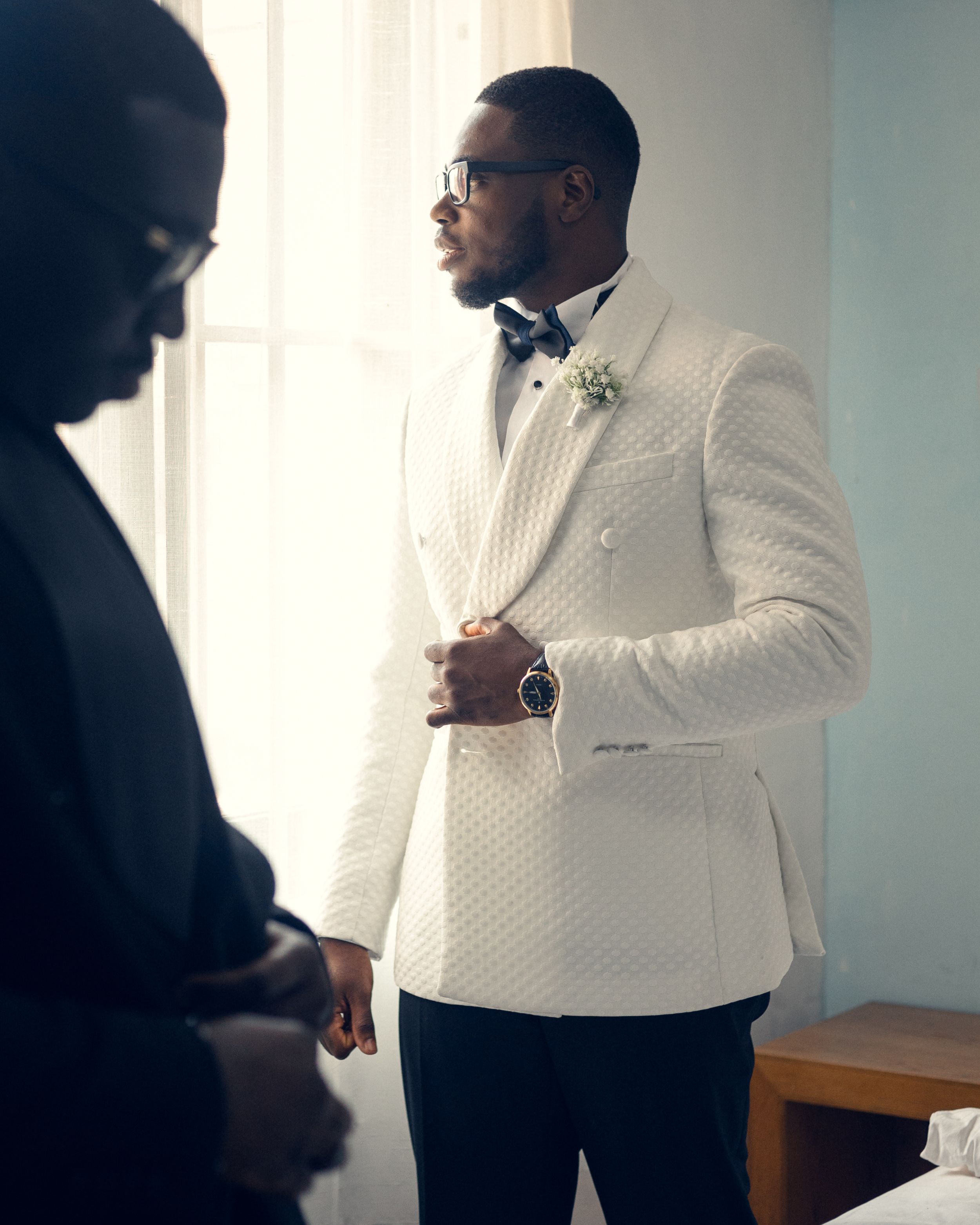 Groom in white quilted tuxedo jacket and bowtie stands in contemplative pose.