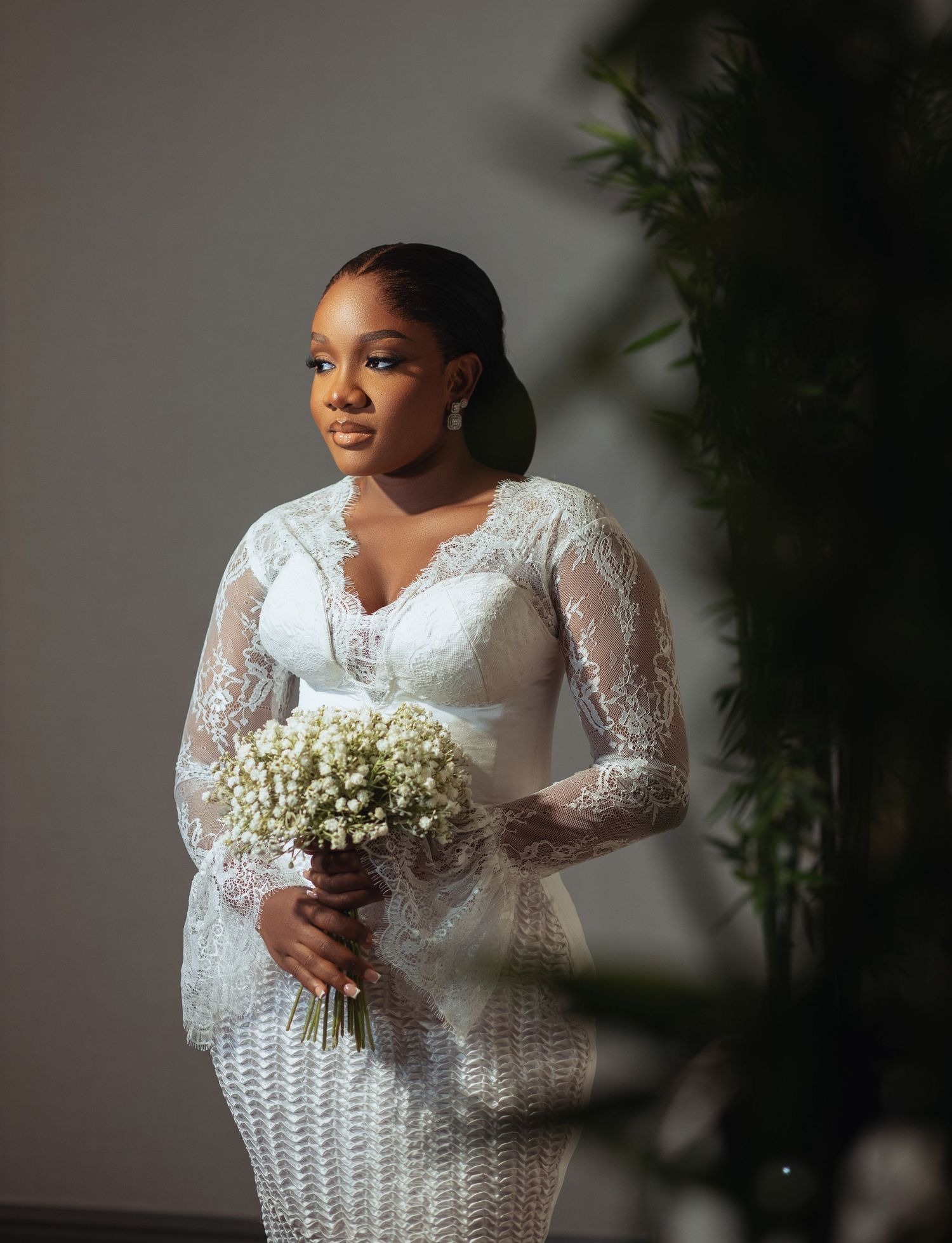 Beautiful bride wearing white lace long sleeve wedding gown holds delicate bouquet of baby's breath flowers against grey wall.