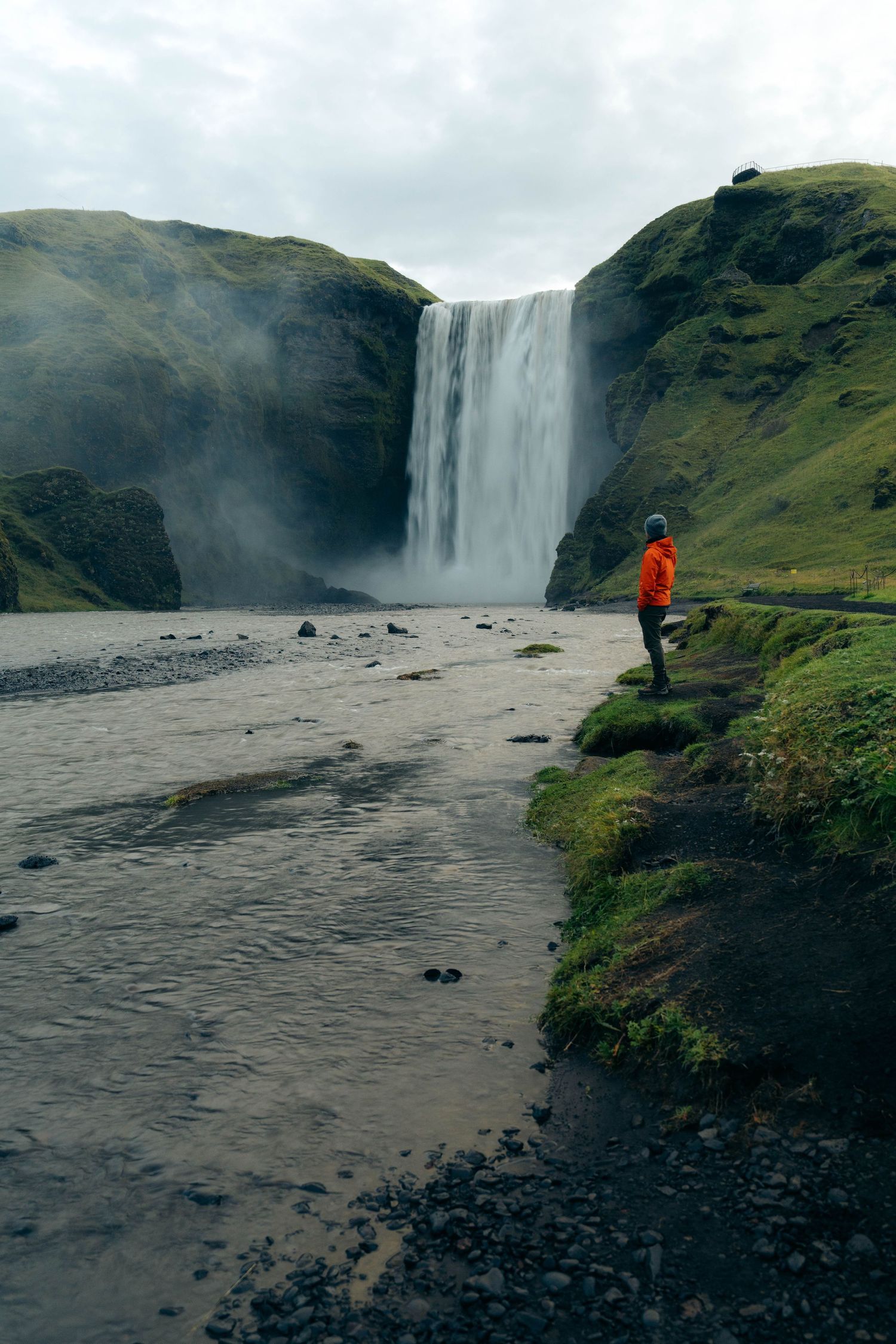 A person in a bright orange jacket stands near the edge of Skogafoss waterfall in Iceland on a misty day.