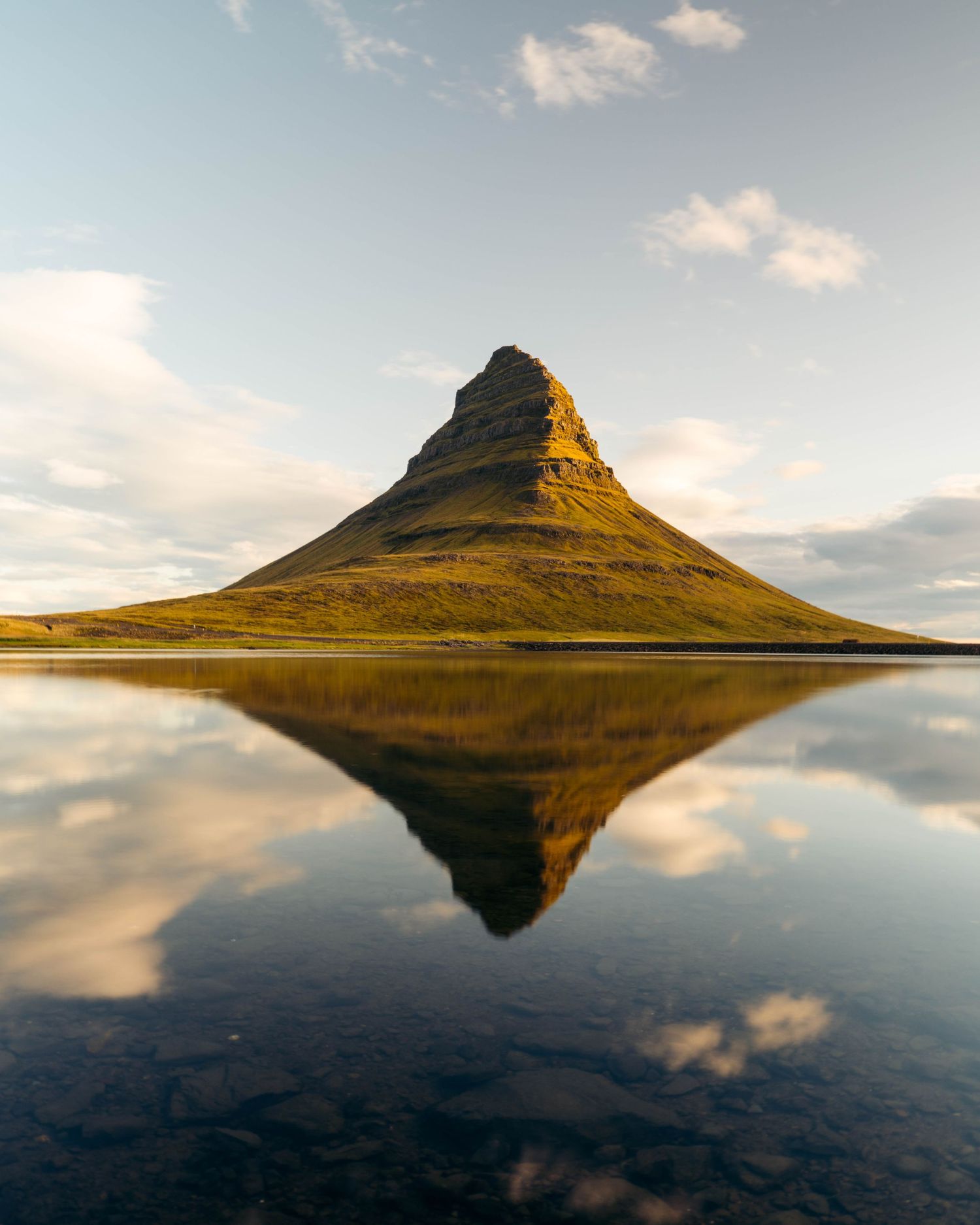 Kirkjufell mountain in Iceland perfectly reflects in calm waters during golden hour with scattered clouds in the sky.