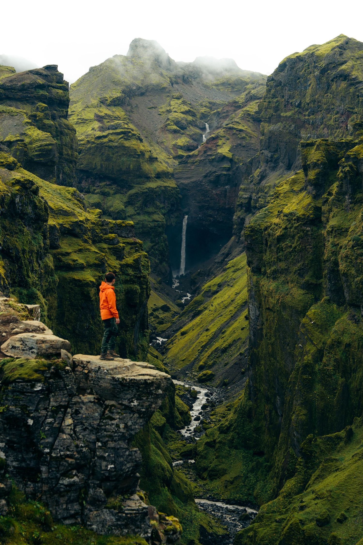 A hiker in an orange jacket stands on a cliff edge overlooking a dramatic waterfall in Iceland's moss-covered mountains.