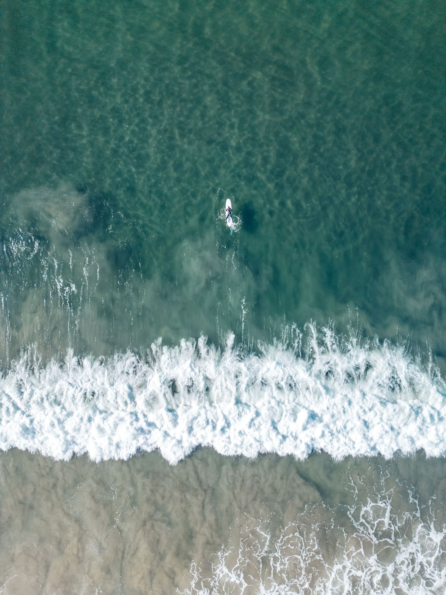 Bird's eye view of a lone surfer in turquoise waters waiting for waves near a white sandy beach.