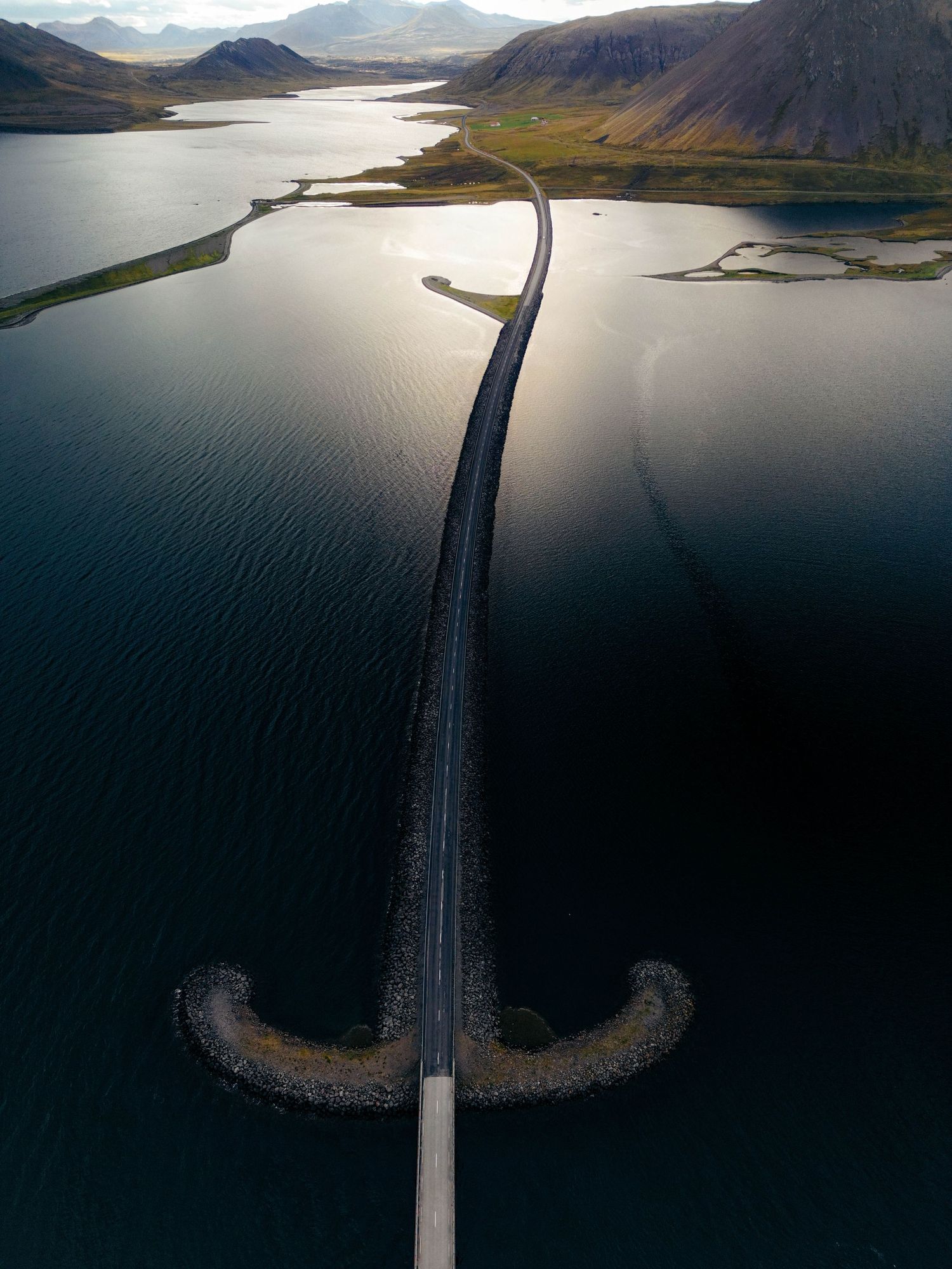 Aerial view of a long winding road bridge crossing a scenic fjord in Iceland with mountains and water at sunset.